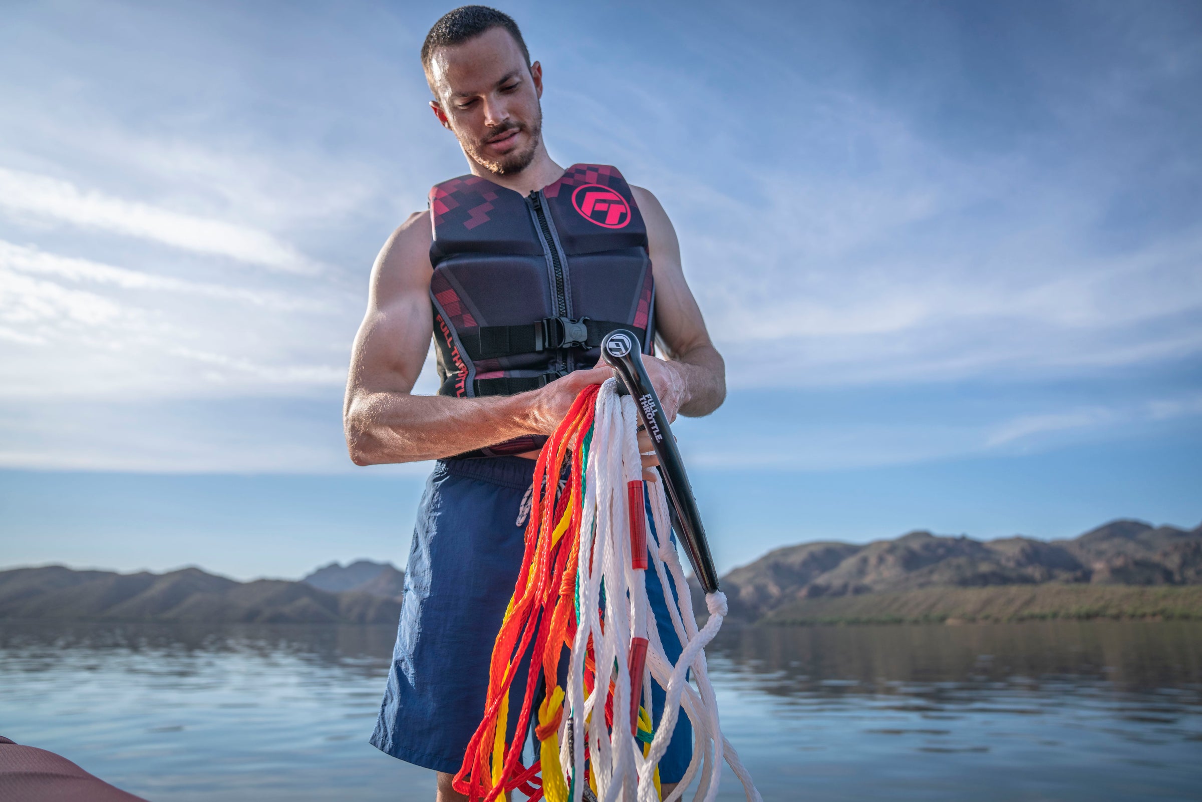 Man in a life vest holds colorful ropes on a boat with mountains and a lake in the background.