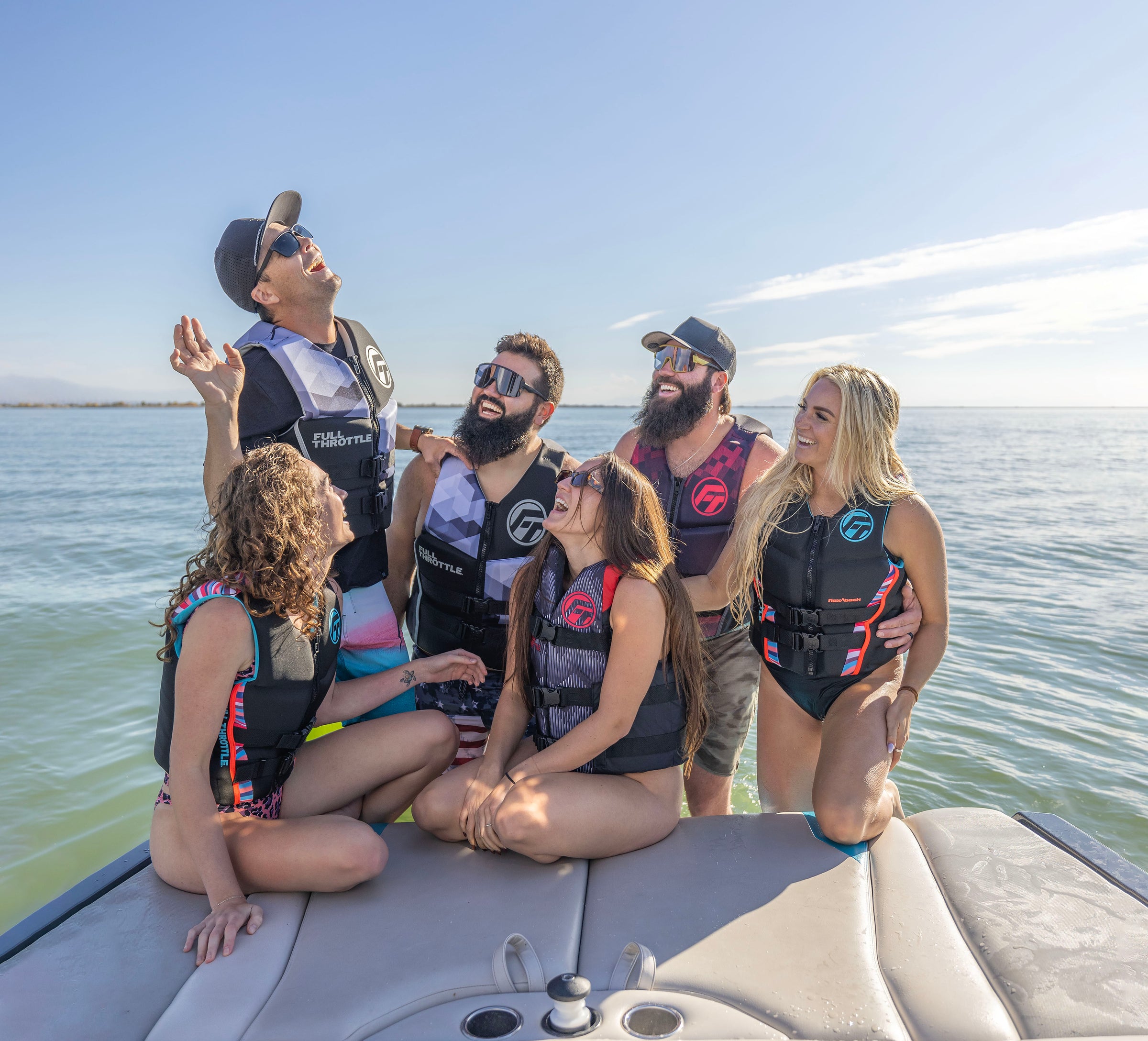 A group of six people in swimsuits and vests laugh and chat on a boat in sunny weather.