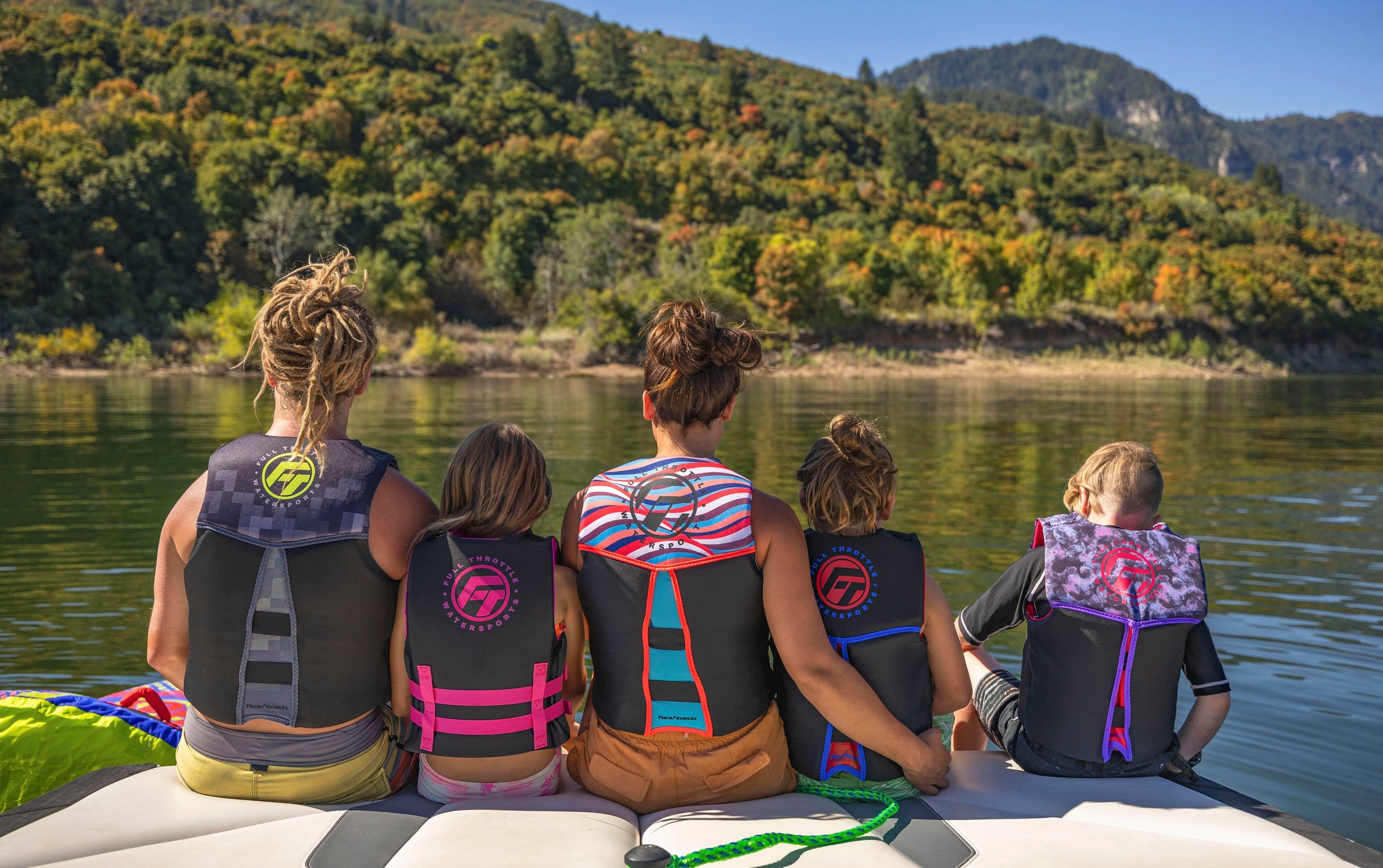 Five people in life vests sit on a boat, facing a scenic view of lush green hills and calm water.