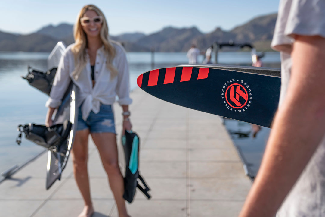 Two people on a dock holding wakeboards, with mountains and a lake in the background.