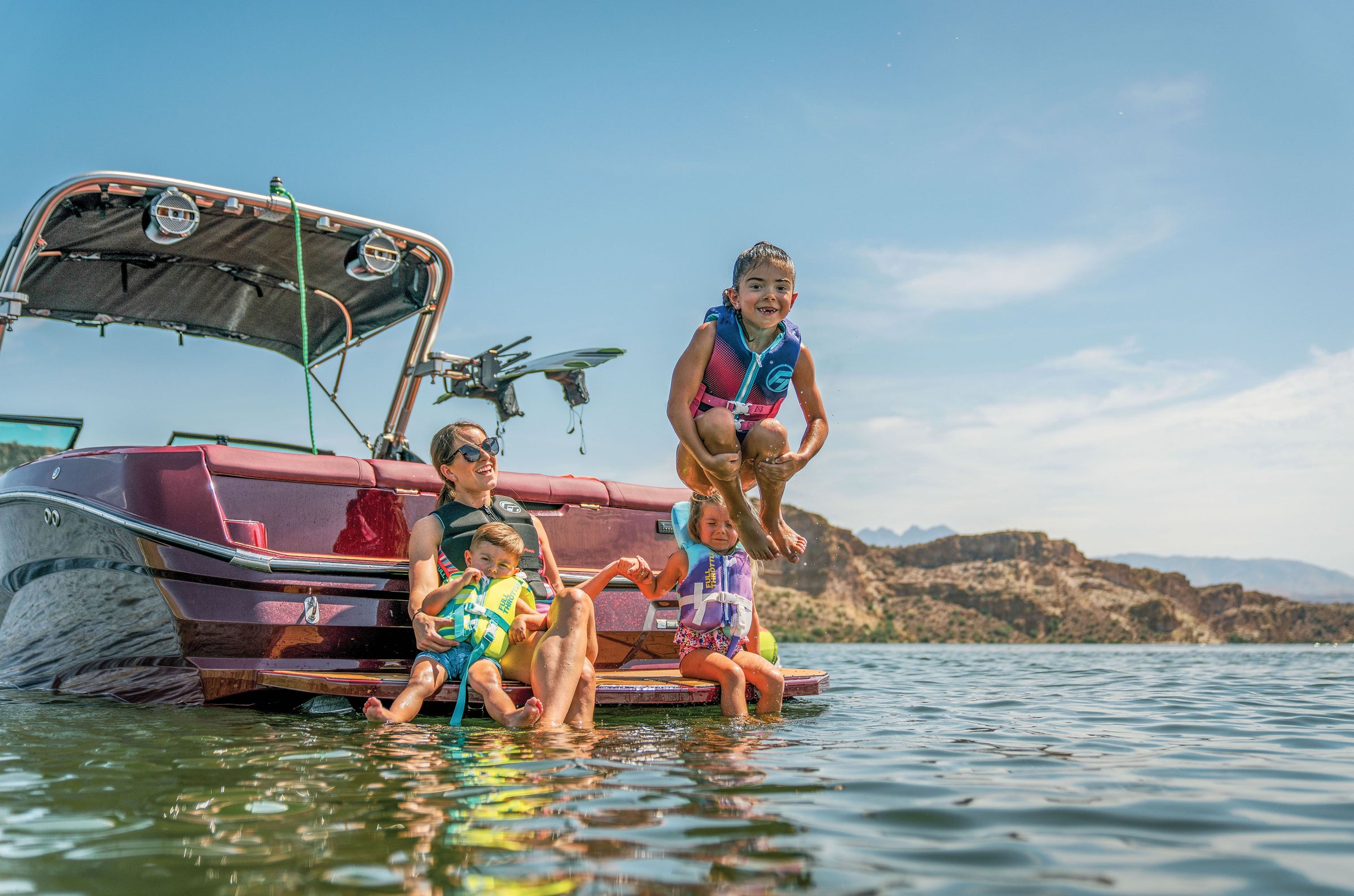 Family sitting on a boats edge in a lake, with a child jumping into the water, surrounded by mountains.