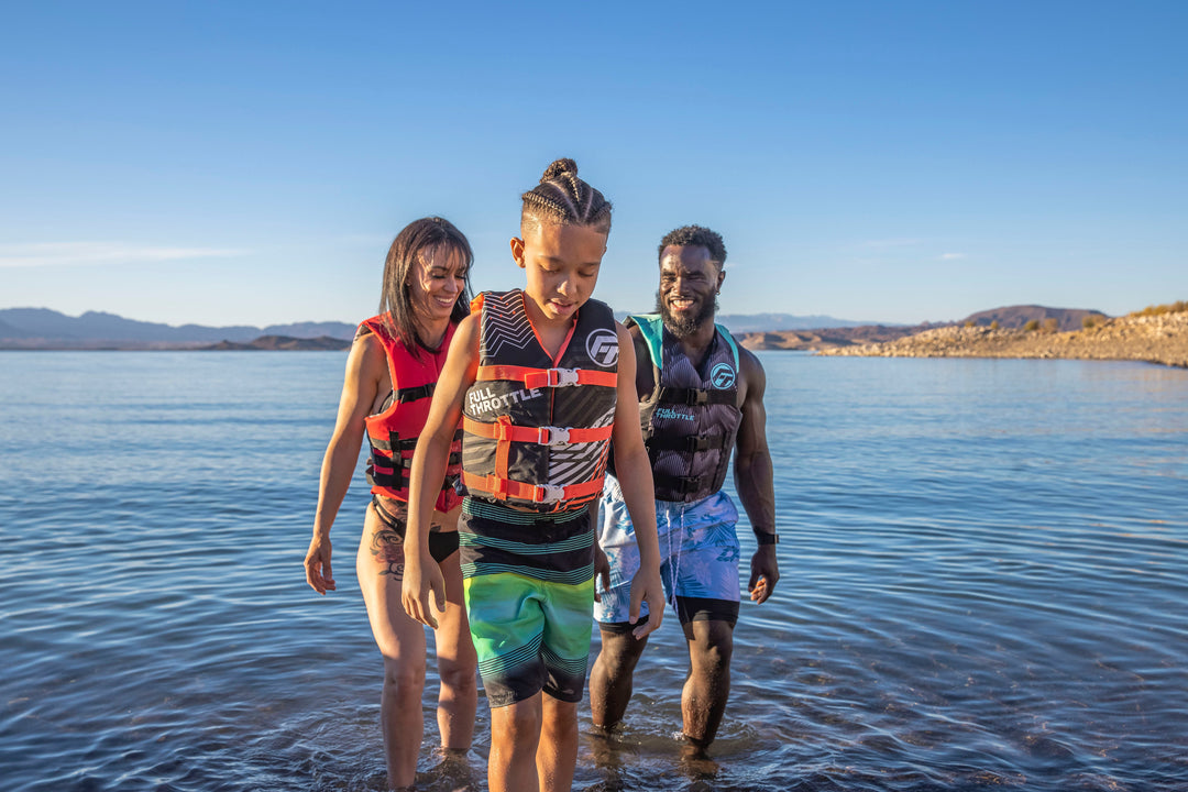 A group of three people in life vests walking in shallow water, smiling under a clear blue sky.