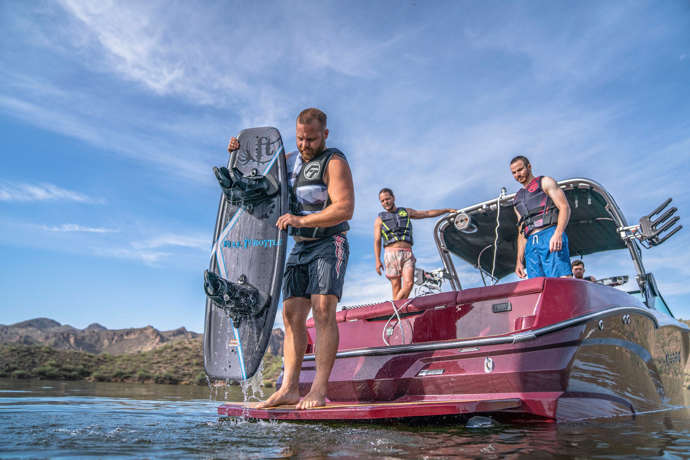 A man with a wakeboard stands on a boat dock while others, in life vests, sit and stand on the boat under a clear sky.
