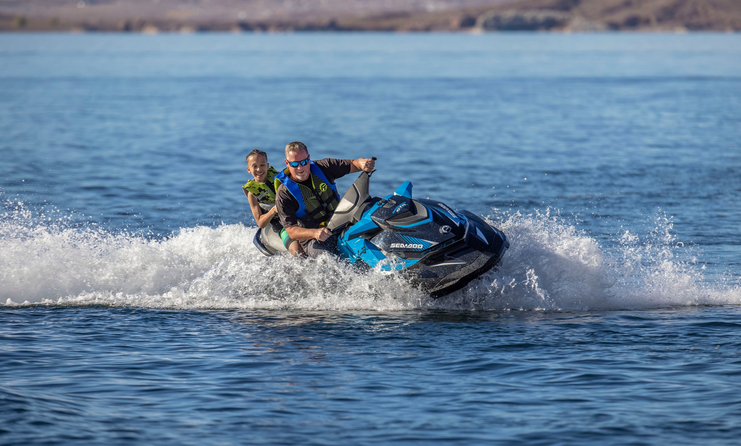 Man and child riding a blue Jet Ski on a lake, creating waves, with mountains in the background.