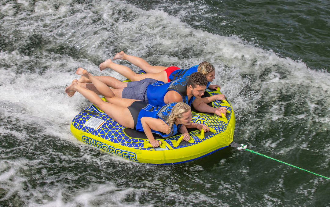 Three people wearing life jackets ride a yellow inflatable tube on water, creating a wake behind them.