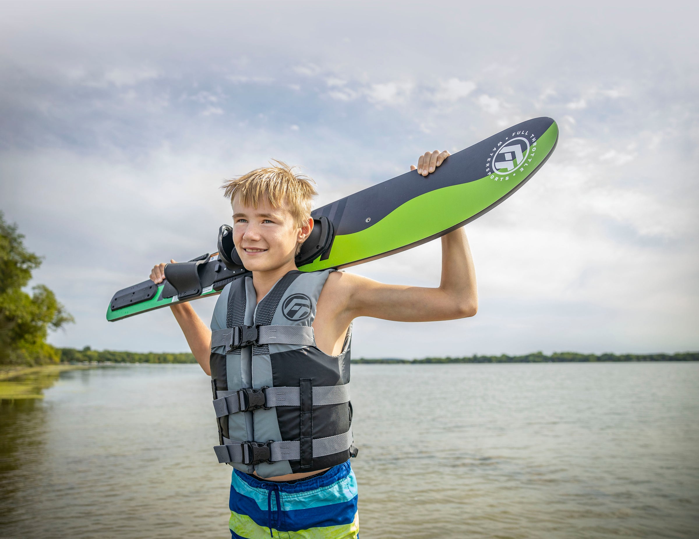 Boy in life jacket holding a green and black wakeboard on his shoulders by a lake on a cloudy day.