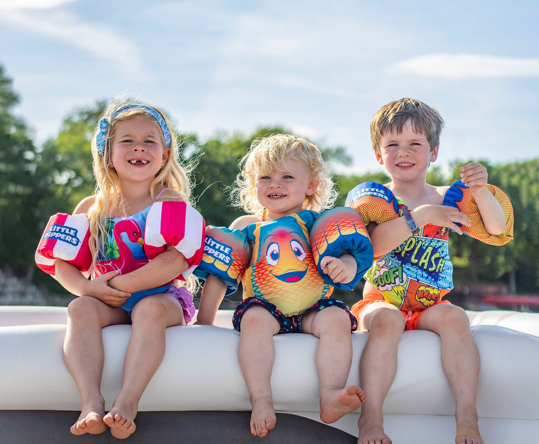 Three children in colorful swimsuits and arm floaties sit on a boat edge, smiling under a sunny blue sky.