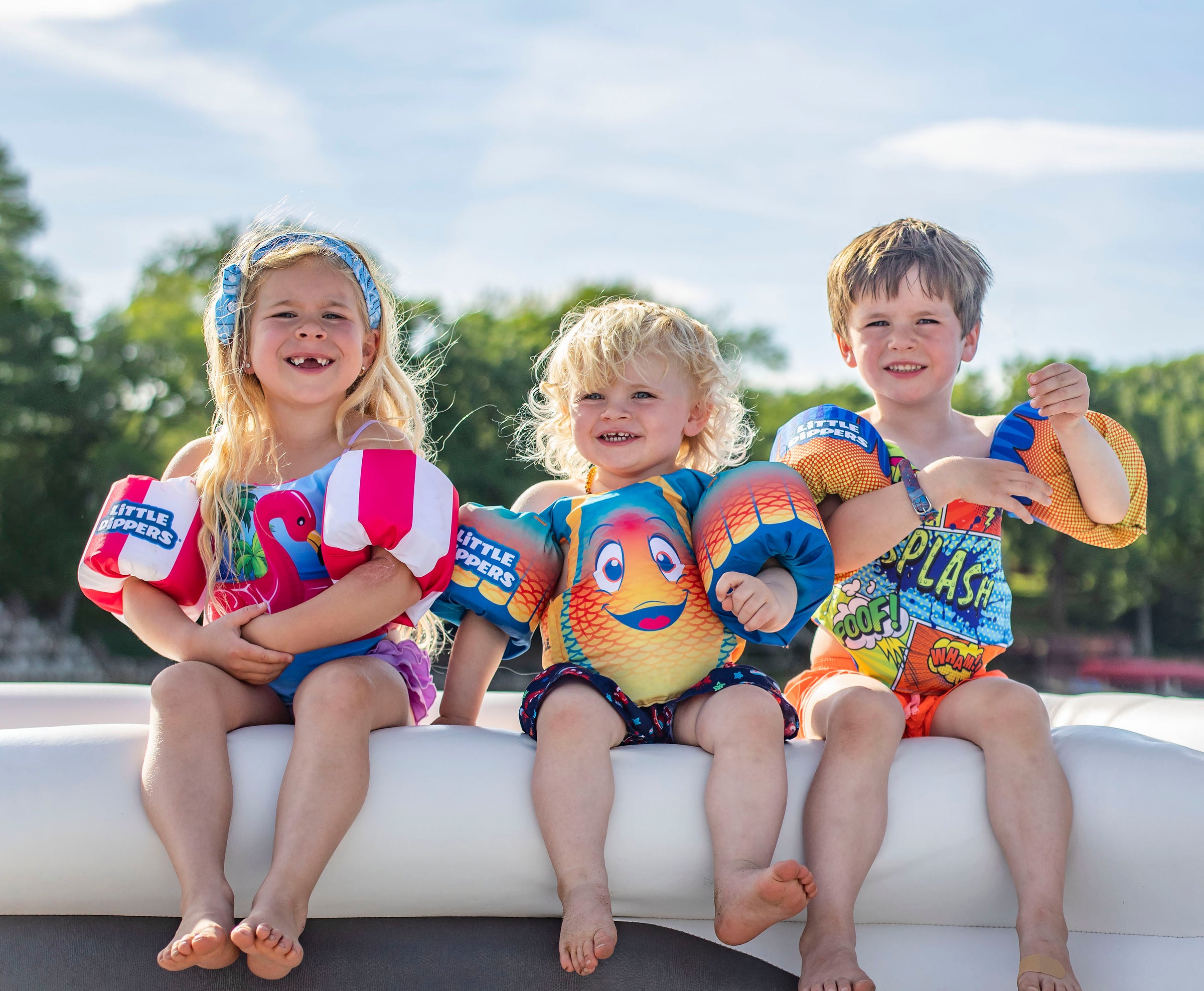 Three children in colorful swimsuits and arm floaties sit on a boat edge, smiling under a sunny blue sky.
