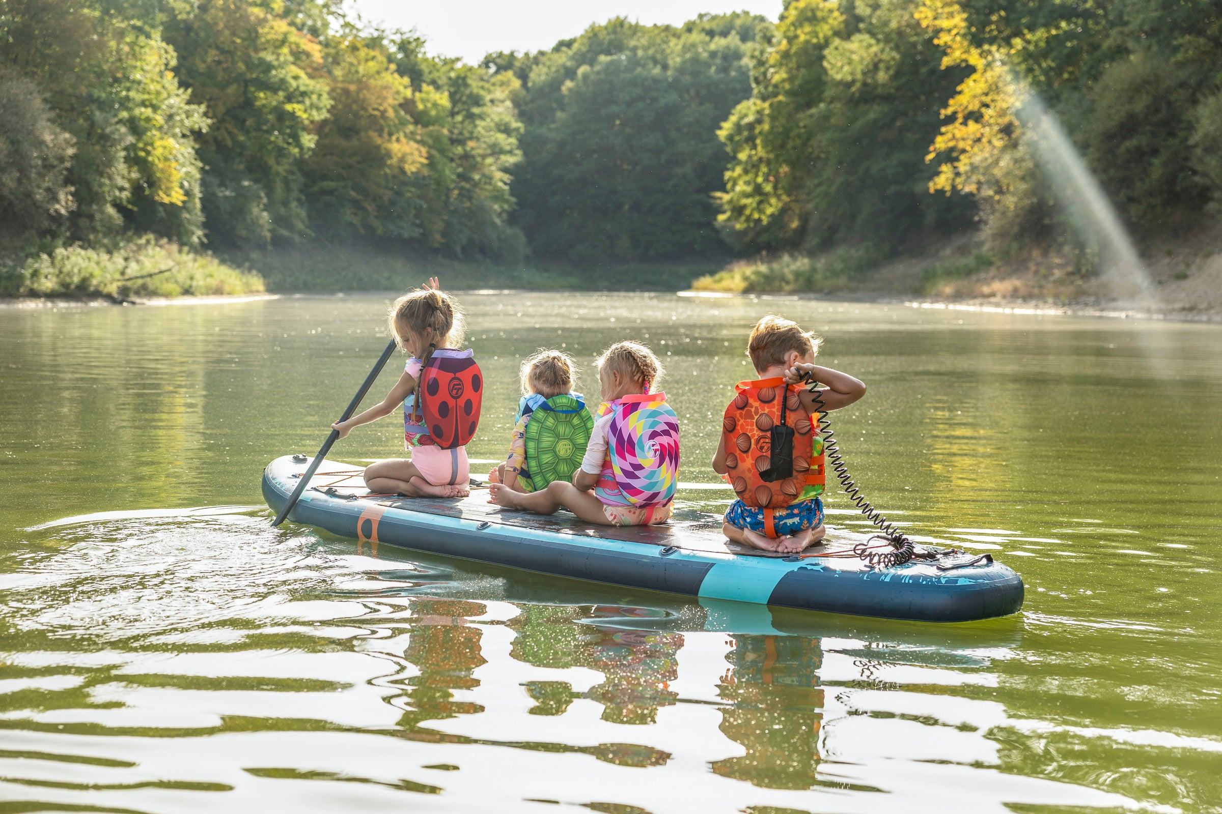 Four children in life jackets sit on a paddleboard on a calm river.