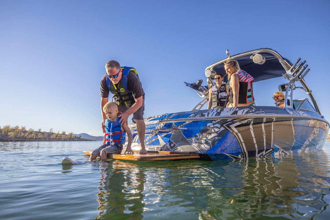 A man helps a child water ski near a blue boat with people onboard.