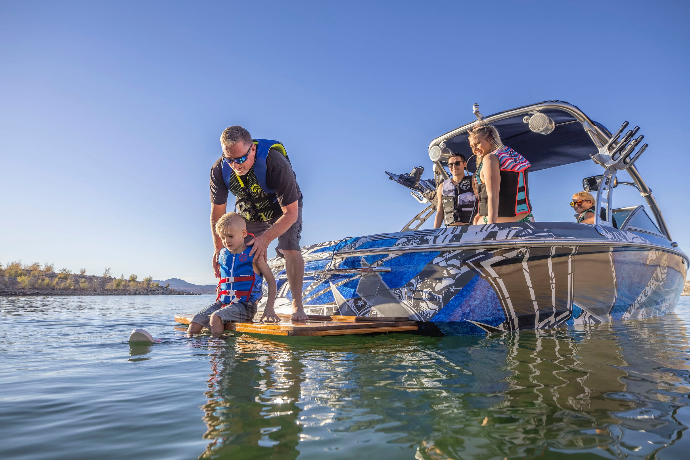 A man helps a child water ski near a blue boat with people onboard.